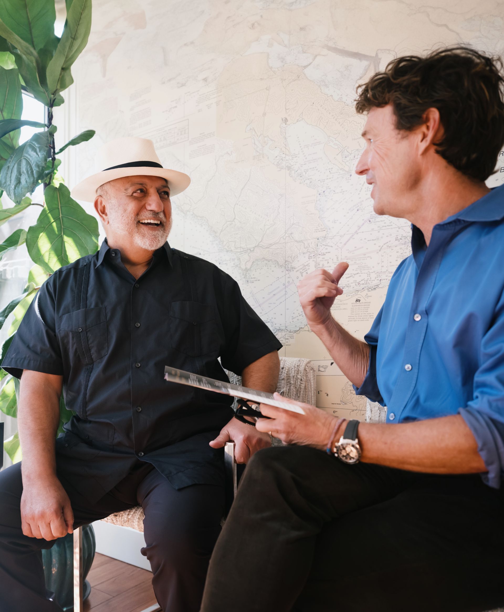 Two men sitting and engaging in a lively conversation indoors with a large map on the wall behind them.